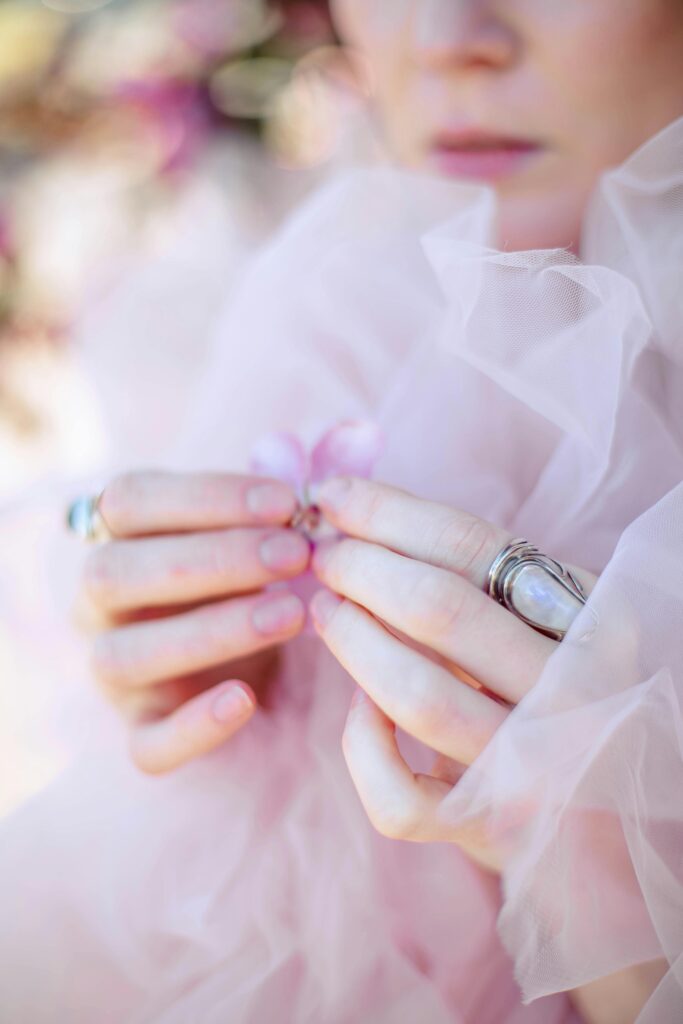 Close-up of a bride's hands holding a pink flower, adorned with rings against a soft pink fabric backdrop.
